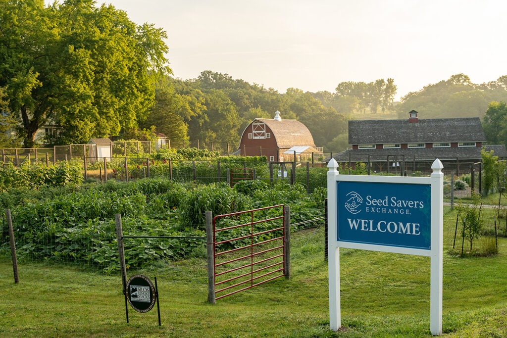 A blue sign, a lush green garden, green trees, a building, and a barn