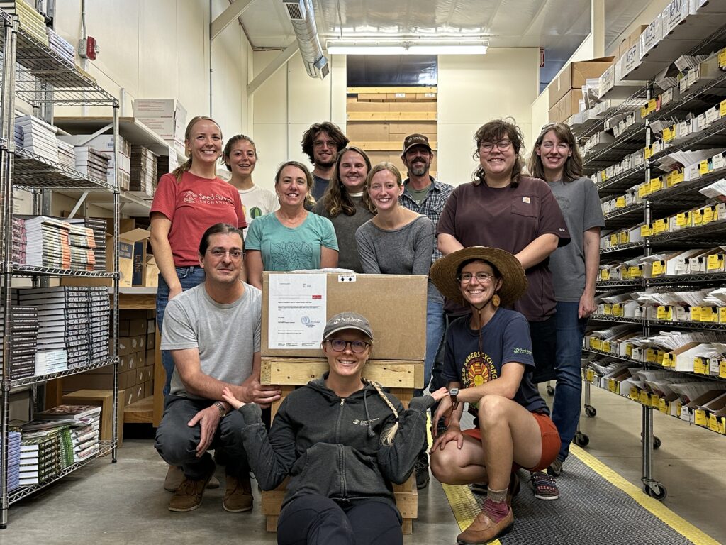 Eleven individuals cluster around a large cardboard box, flanked by shelves of seeds.