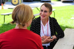 A woman in a red shirt sits at a picnic table with her back to the camera and converses with a woman in a white shirt with a black blazer