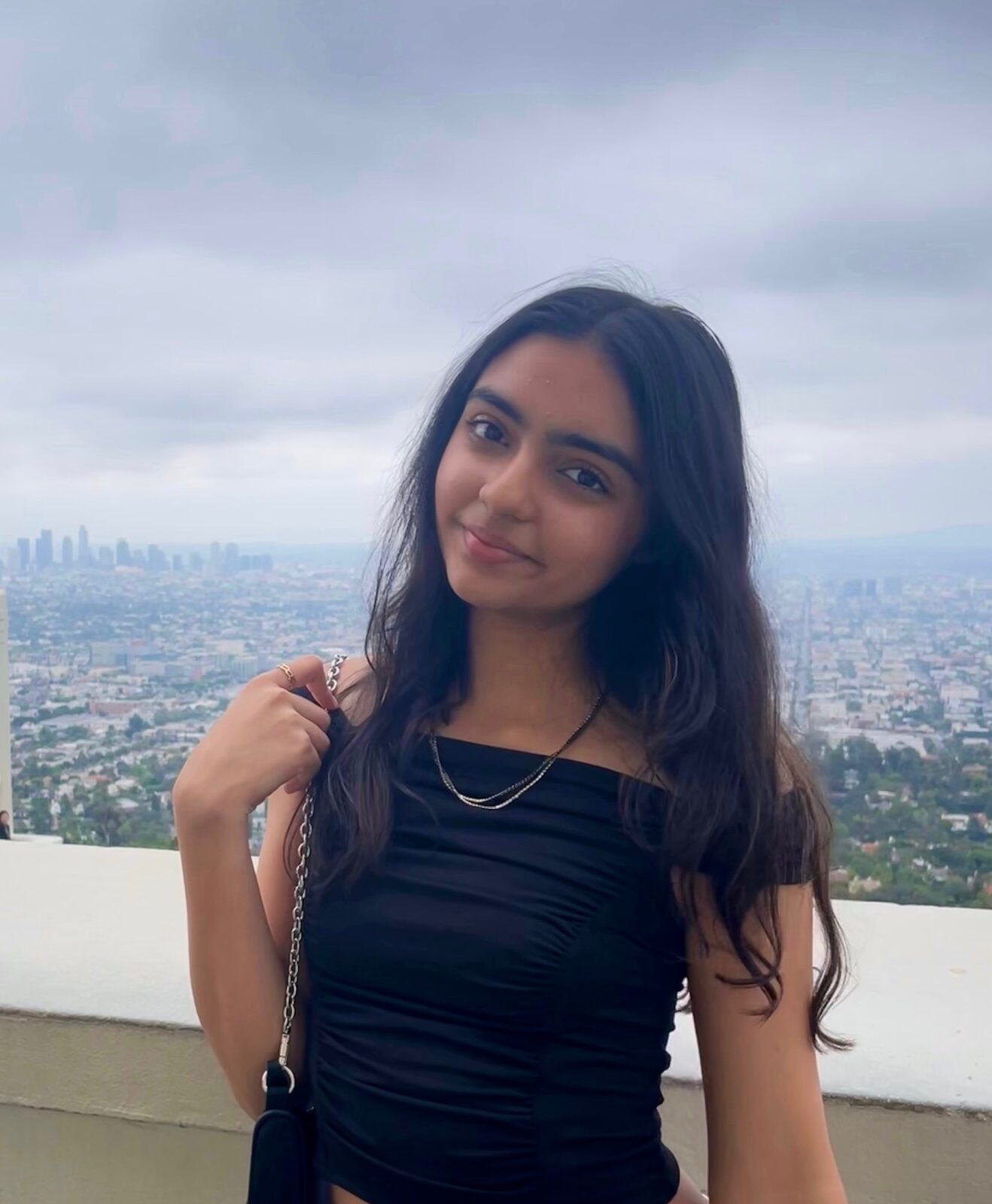 A young woman with long hair looks to camera with a cityscape behind her.