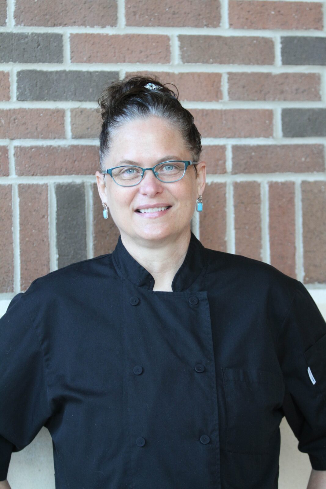 A woman in a black chef's jacket and glasses smiles to camera.