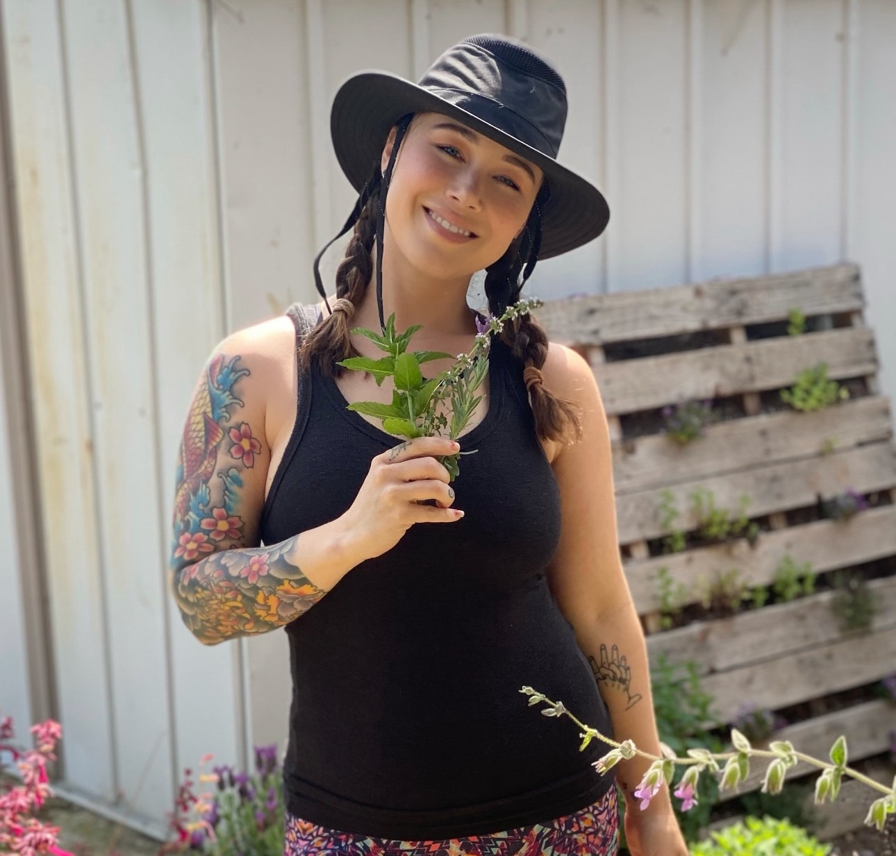Woman with a wide brim hat smiles to camera and holds flowers by her chest.