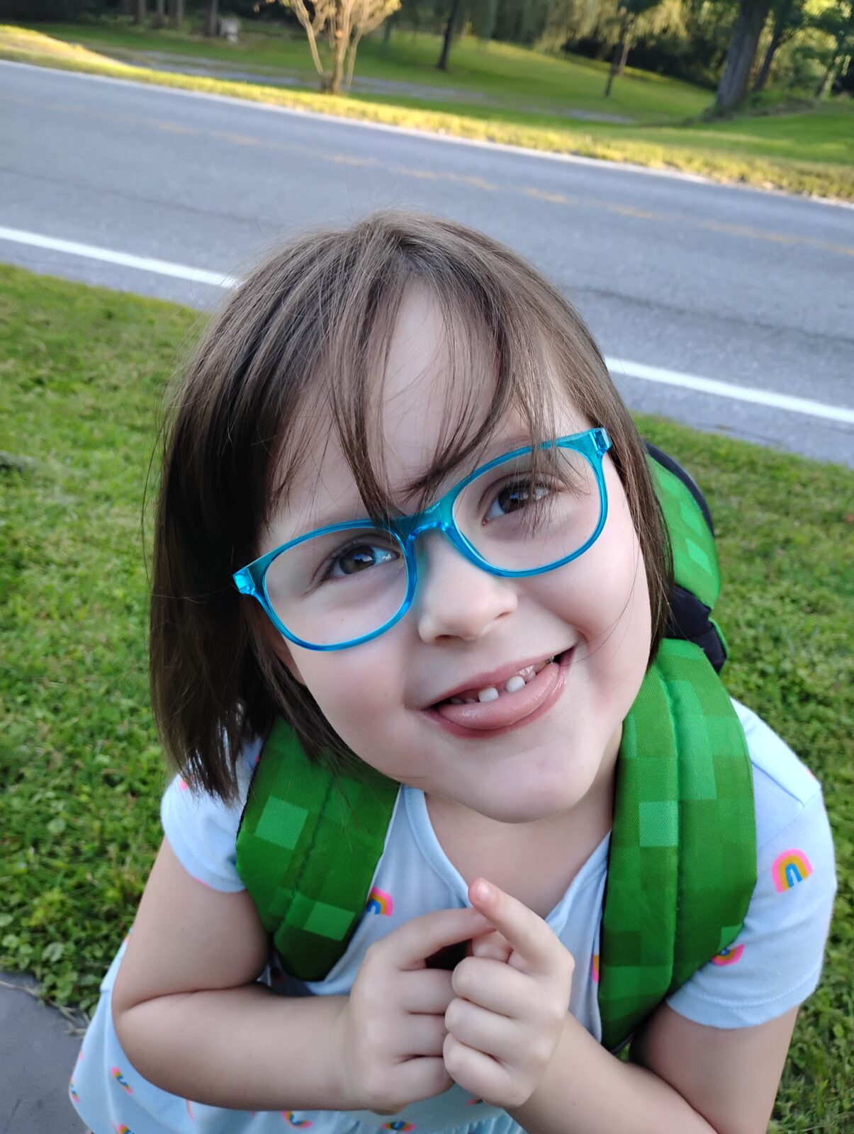 Young child with glasses wearing a backpack smiling and sticking her tongue out at the camera.