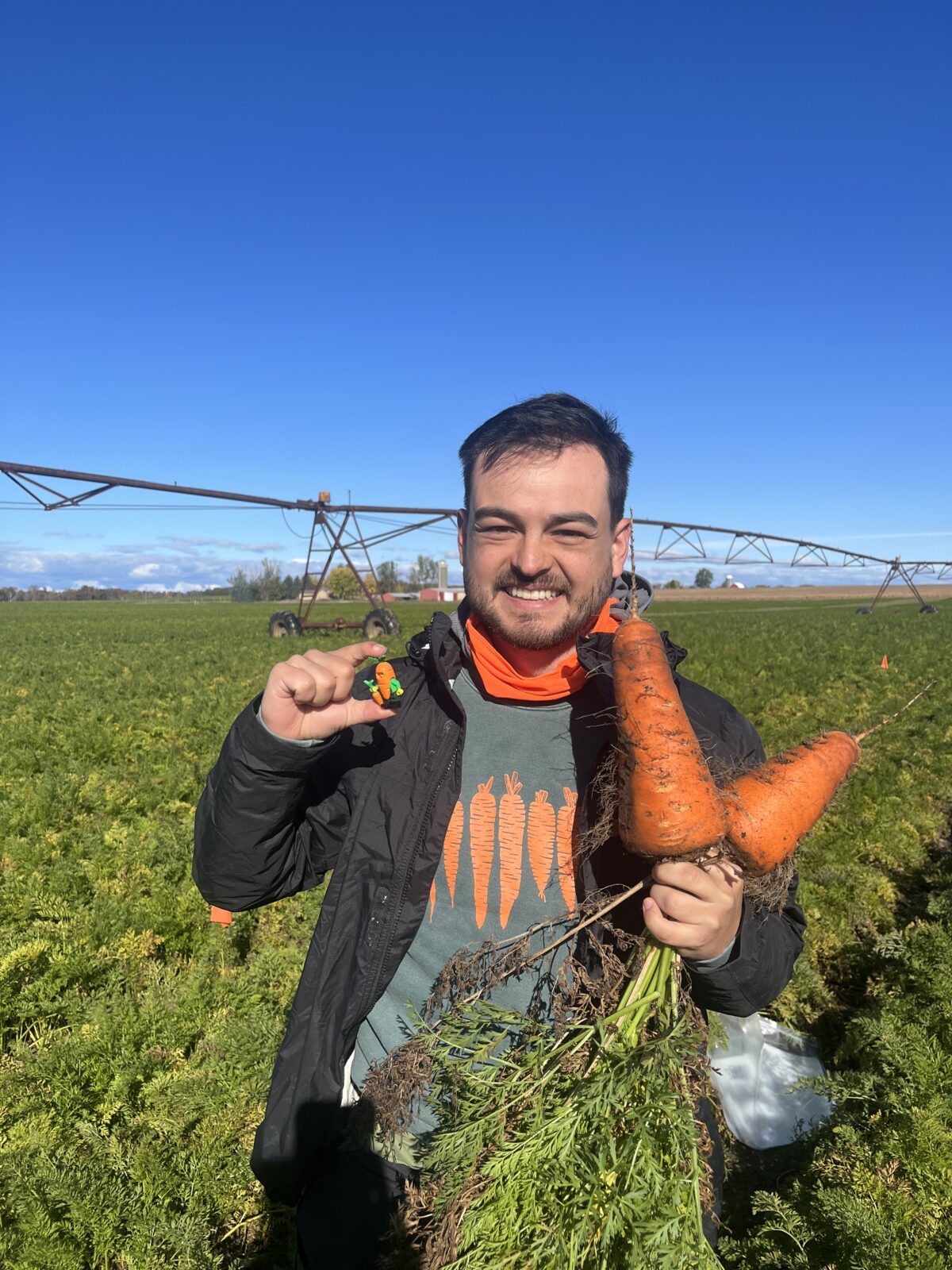 A man stands in a field holding two large carrots, and a small toy, smiles to camera.