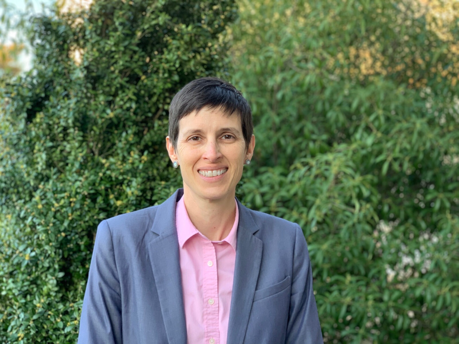 A woman in a gray blazer smiles to camera standing in front of greenery.