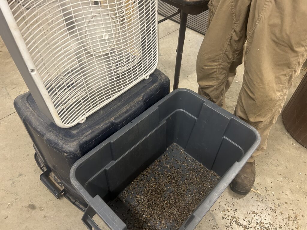 A box fan on top of an overturned bucket with a bucket of seeds in front of it