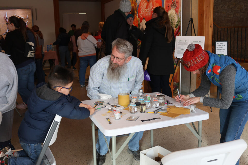 Man with a beard sits at a card table talking to a young child looking at the seeds in front of him.