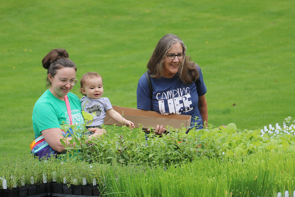 Two women, one is holding a baby, look over pallets of heirloom transplants.
