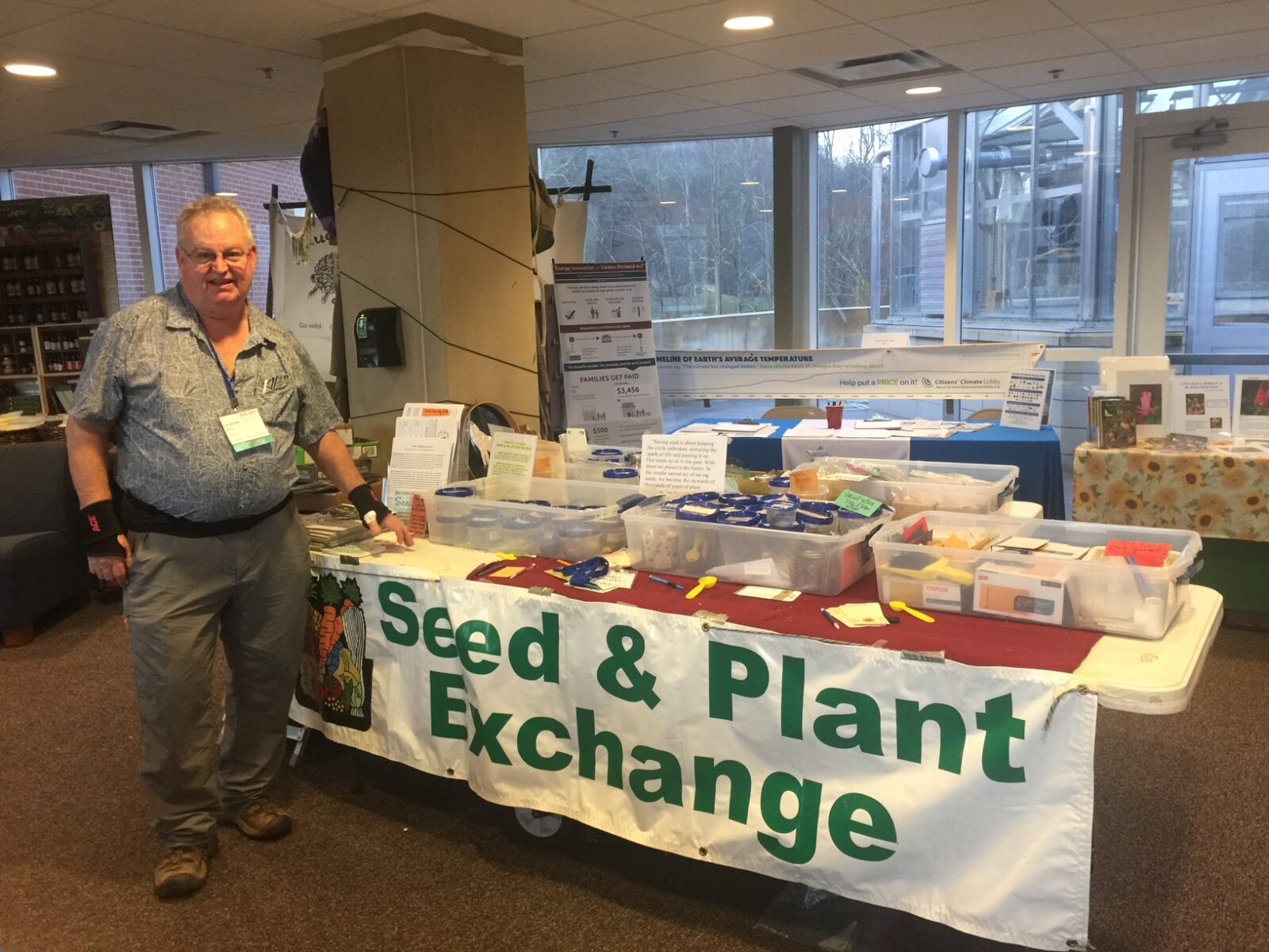 older man standing next to a long table with seeds on it and a sign reading 