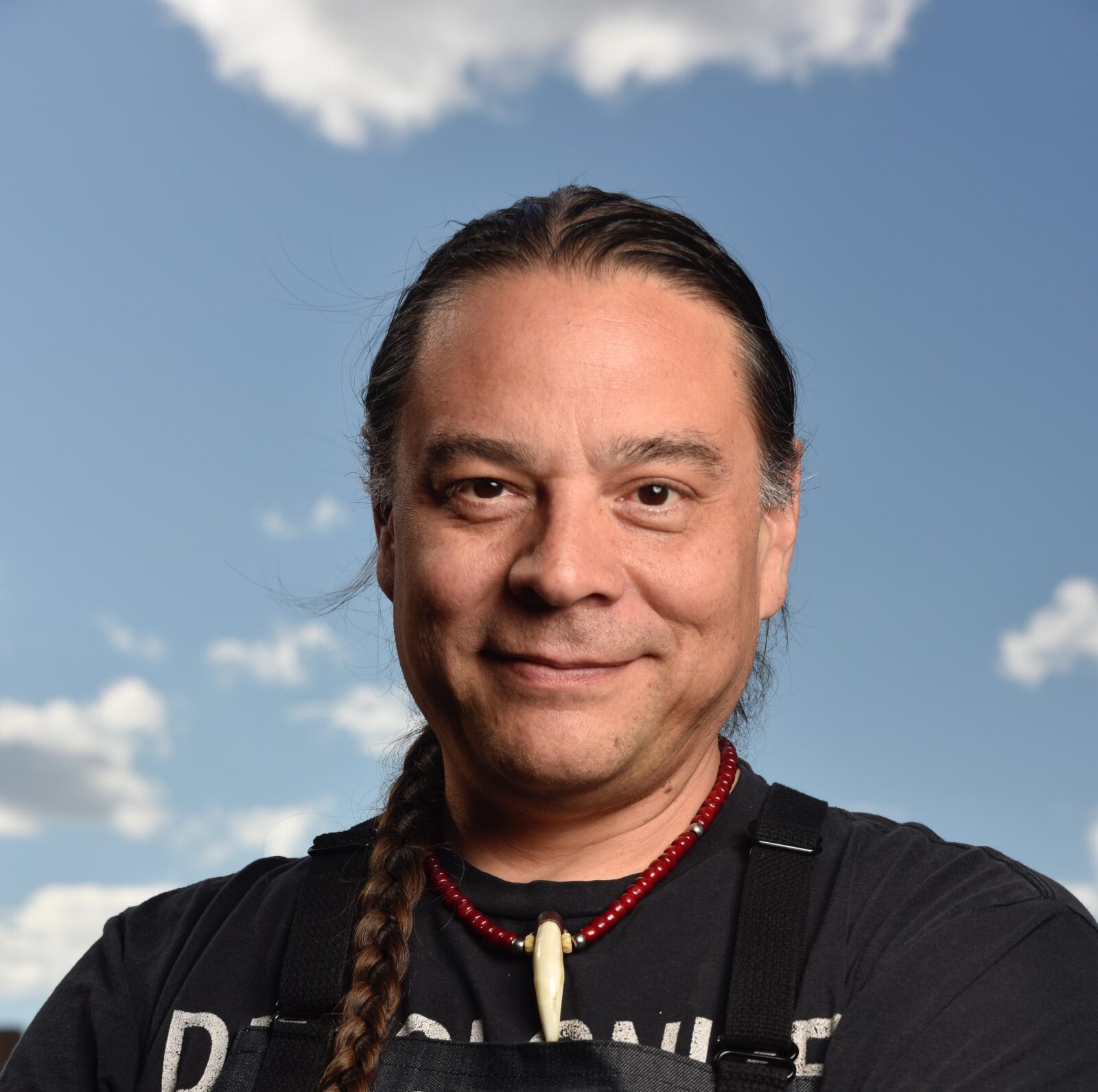dark-haired man with long braids, black shirt, and Owamni apron, against background of blue sky with clouds